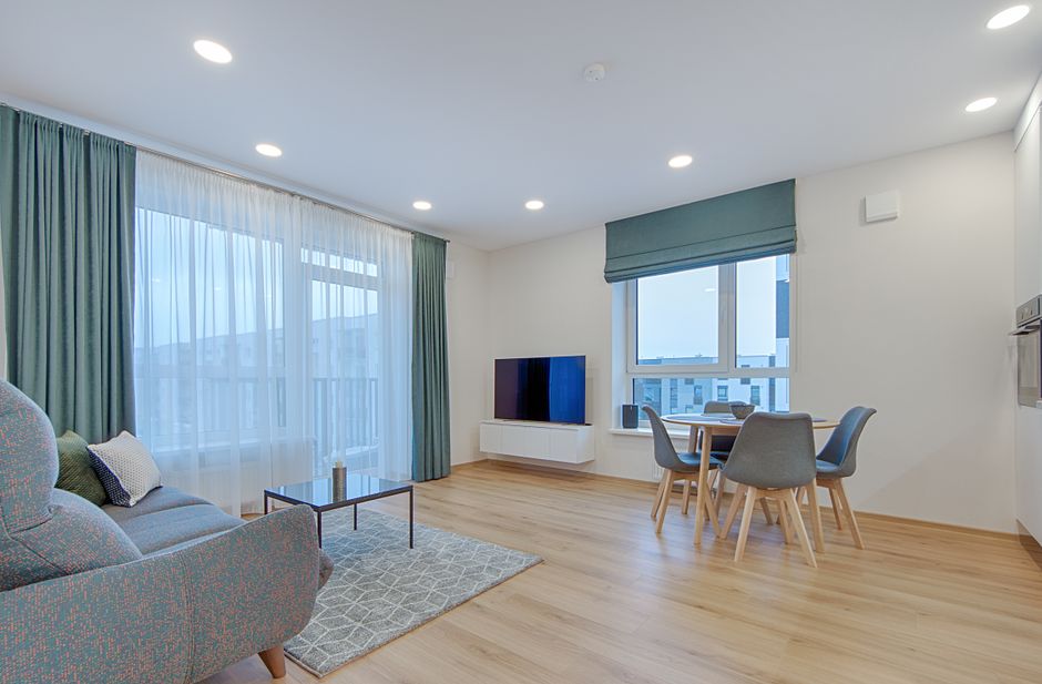 A modern, bright living room with light wooden flooring and white walls illuminated by recessed ceiling lights. A large window with sheer white curtains and teal drapes allows natural light to fill the space, while another window has a teal roller blind. The room features a patterned upholstered sofa with a cushion, a small coffee table, and a flat-screen TV mounted on a white floating cabinet. The dining area includes a round wooden table with four upholstered chairs in light grey. The surfaces are clean and well-maintained, reflecting a professional domestic cleaning service as suggested in the SW19 upholstery and rug cleaning guide for flats by Carpet Cleaning Wimbledon.