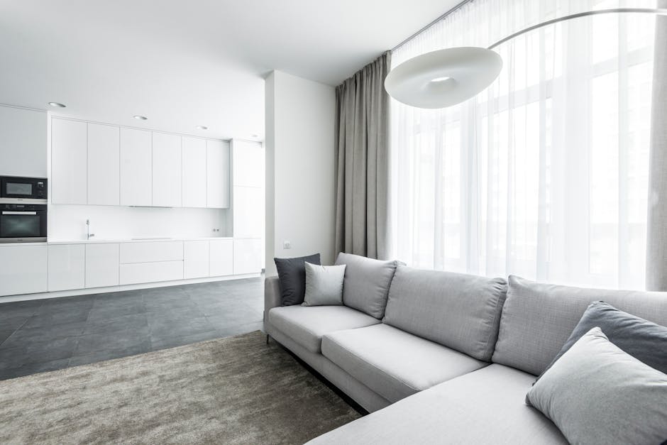 A bright, modern living room with a large light gray fabric sofa adorned with matching cushions, positioned on a neutral-toned rug. The room is illuminated by natural light streaming through sheer white curtains covering floor-to-ceiling windows. Adjacent to the living space, an open-plan kitchen features sleek white cabinetry, built-in appliances, and a minimalist design. The dark tiled floor contrasts with the white surfaces, creating a clean and tidy appearance. The overall scene emphasizes an atmosphere of freshness and cleanliness, consistent with professional domestic surface cleaning and maintenance, as provided by Carpet Cleaning Wimbledon, showcased within the SW19 upholstery and rug cleaning guide for flats.