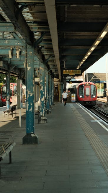 The image shows a clean, well-maintained train station platform with a concrete surface and tactile paving near the edge for safety. Blue metal columns support an overhead canopy with visible structur