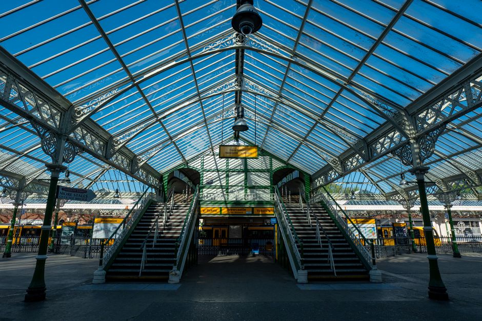 The image shows a clean, well-maintained train station platform with a concrete surface and tactile paving near the edge for safety. Blue metal columns support an overhead canopy with visible structur