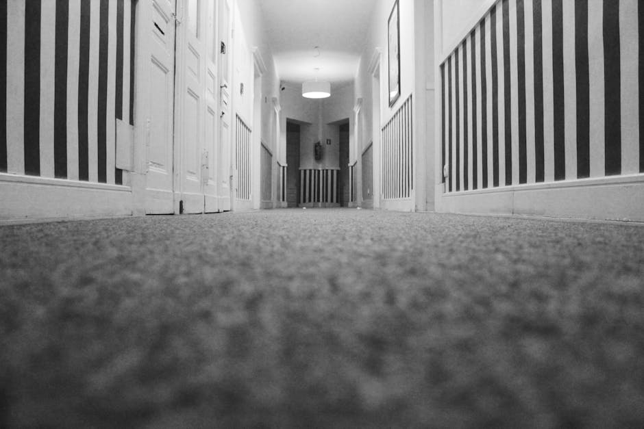 A black and white photograph of a residential hallway viewed from a low angle, showing a textured carpet floor with a subtle pattern. The hallway has white wooden doors on the left side and a striped wallpaper pattern on the right wall, featuring alternating dark and light vertical stripes. The ceiling is illuminated by a centrally placed round light fixture, casting soft light across the space. The walls and baseboards appear clean and well-maintained. The scene depicts a tidy and hygienic environment, emphasizing surface cleanliness suitable for deep cleaning and sanitisation, aligning with the services offered by Carpet Cleaning Wimbledon for homeowners' surface cleaning needs.