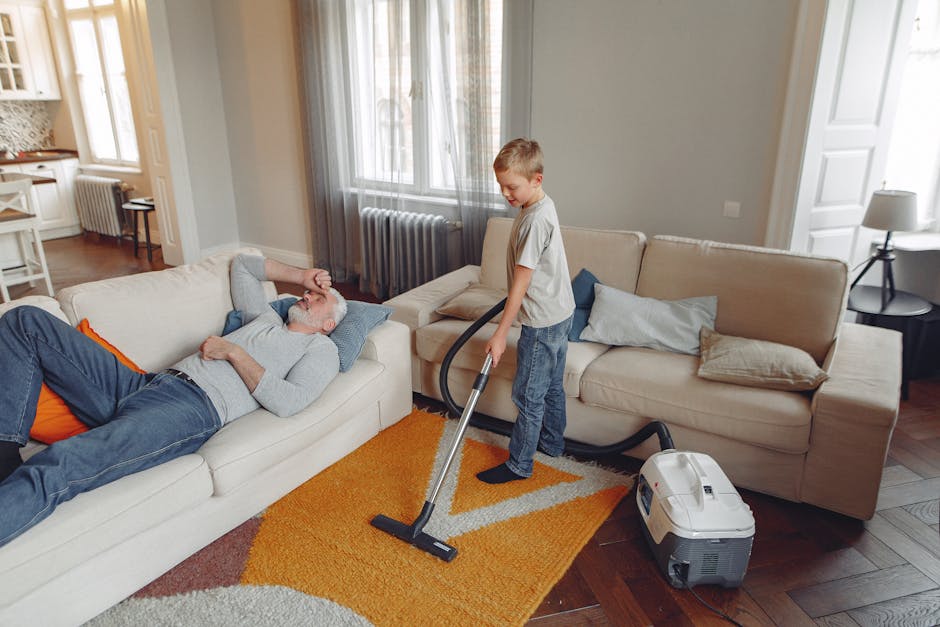 A young boy operating a vacuum cleaner on a small orange and beige rug in a well-lit, modern living room with wooden flooring, a beige sofa with multiple cushions, and large windows with sheer curtains. An older man is lying on the sofa, appearing relaxed. The room features neutral-colored walls, a side table with a lamp, and open doorways leading to other parts of the house. The scene depicts domestic cleaning and surface sanitisation, highlighting deep cleaning efforts by Carpet Cleaning Wimbledon, aligned with their Wimbledon Village carpet cleaning guide for homeowners.